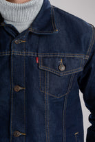 Primer plano del botón metálico grabado y la costura ocre en contraste sobre el denim de una chaqueta de jean azul industrial.