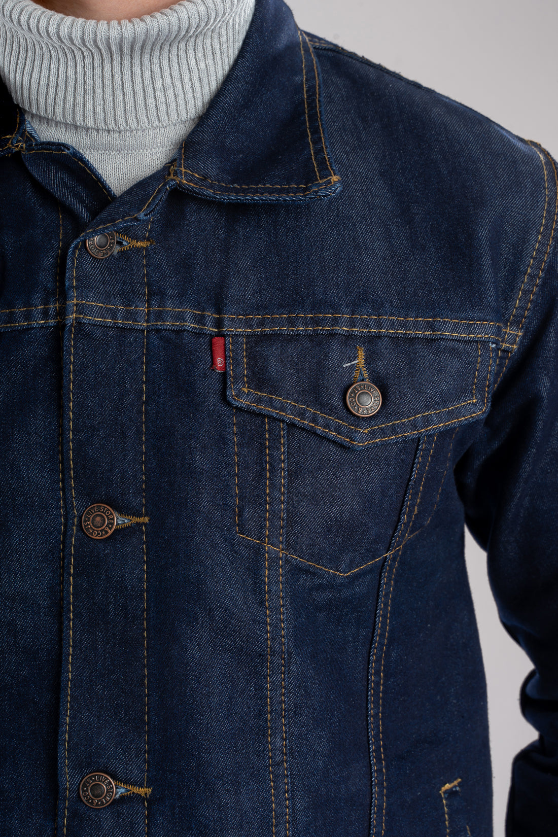 Primer plano del botón metálico grabado y la costura ocre en contraste sobre el denim de una chaqueta de jean azul industrial.