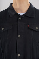 Primer plano del botón metálico grabado y la costura ocre en contraste sobre el denim de una chaqueta de jean negra.